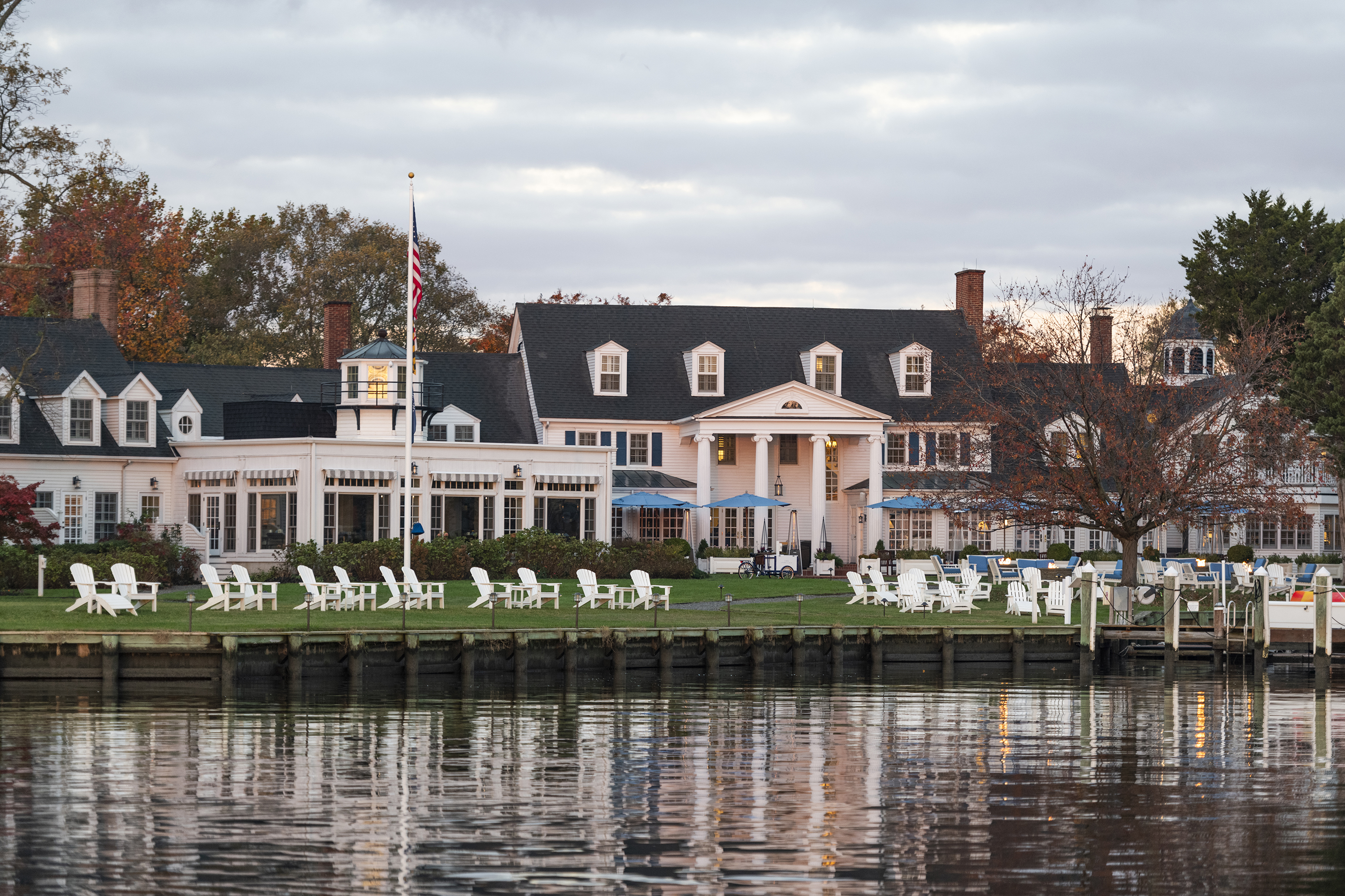 The image shows a large waterfront building with a dock, surrounded by trees, chairs, and an American flag on the lawn.