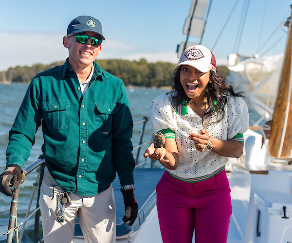 Two people are on a boat, smiling. One is holding an oyster, and they seem to be having a fun time on the water.