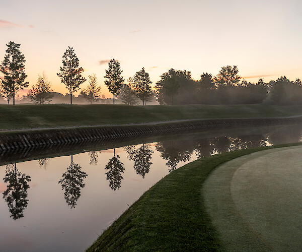 A serene landscape of a golf course at sunrise, with trees reflecting on a calm river.