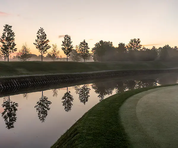 A serene landscape of a golf course at sunrise, with trees reflecting on a calm river.