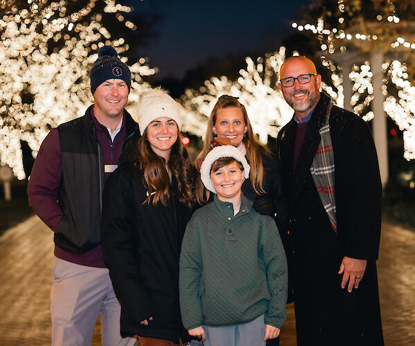 A group of people smiling outdoors at night, surrounded by trees adorned with festive lights.