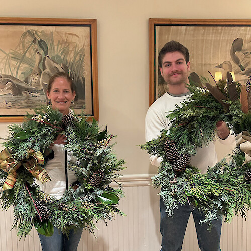 Two people are holding decorative wreaths made of greenery, pinecones, and ribbons. Duck paintings are visible in the background.