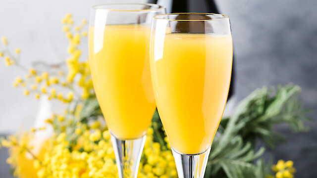 Two glasses of orange juice in flutes with a bottle and yellow flowers in the background.