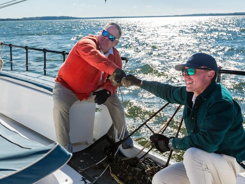 Two people on a boat are having a friendly moment, possibly sharing seafood, with the ocean in the background.