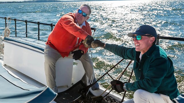 Two people on a boat are having a friendly moment, possibly sharing seafood, with the ocean in the background.