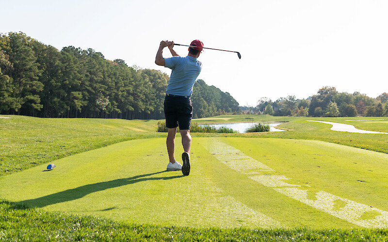 A person on a golf course follows through after a golf swing on the fairway, with trees and a pond in the background.