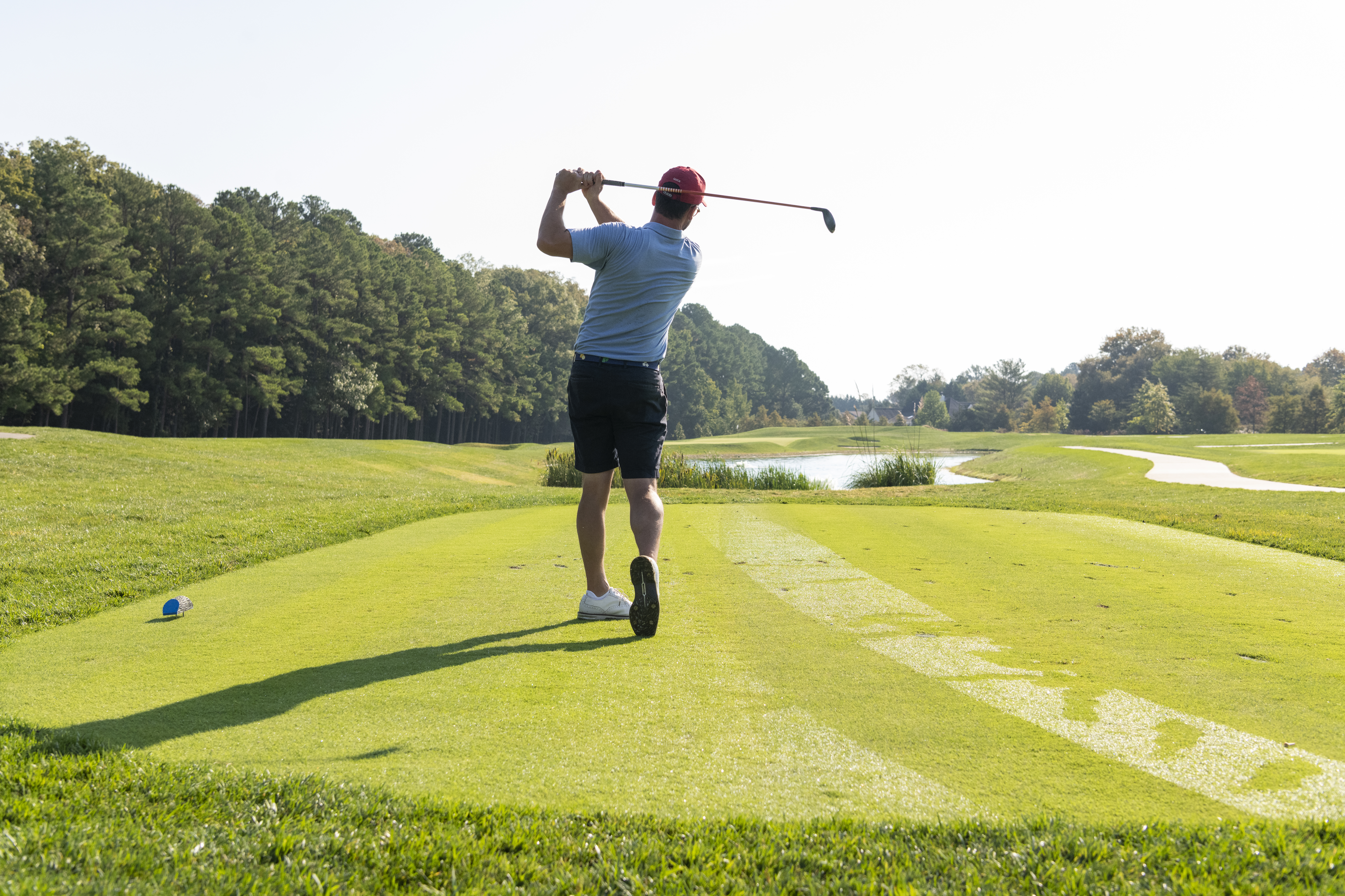 A person is playing golf, hitting a ball on a lush green course, surrounded by trees and a pond in the background.
