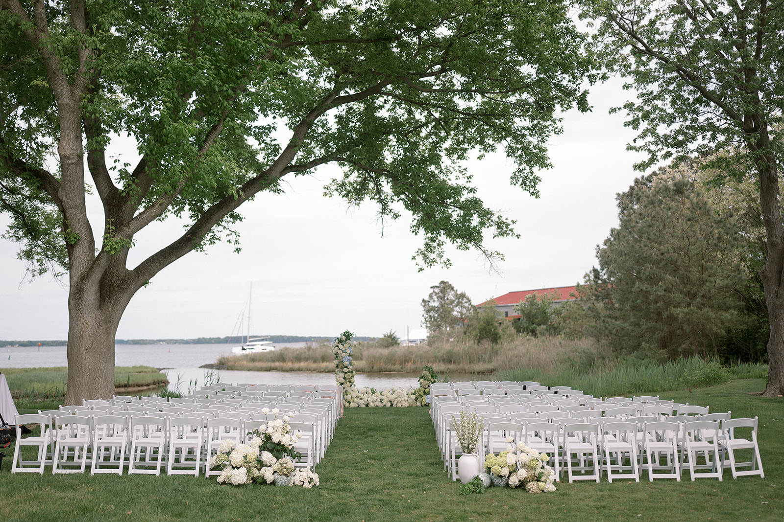 An outdoor wedding setup with white chairs and floral arrangements, under trees beside a body of water.