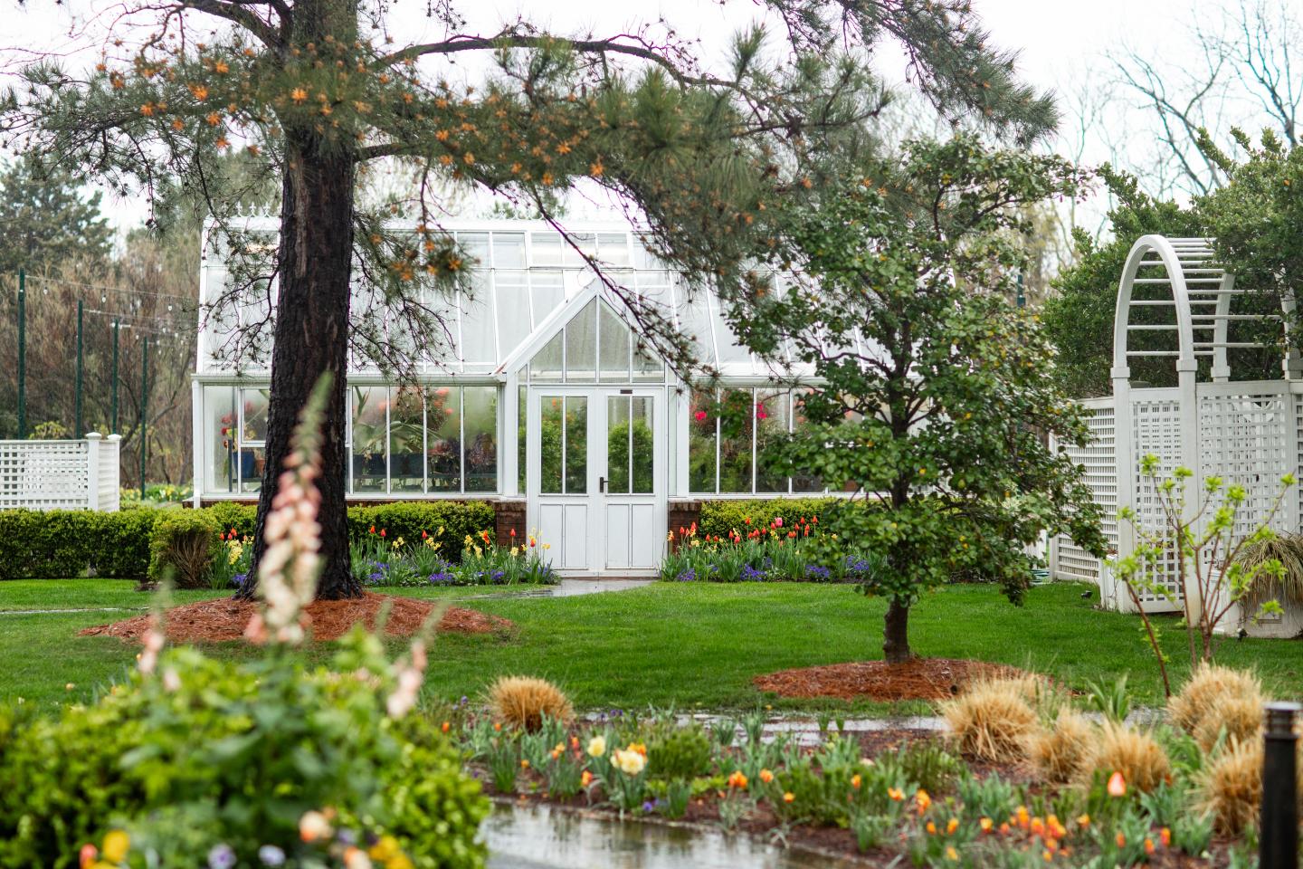 White greenhouse surrounded by lush garden and colorful flowers.